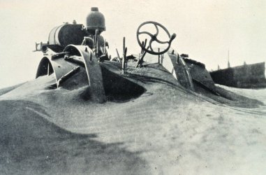 Machinery covered by sand, date unknown An old black and white photograph of indistinct machinery with levers and a steering wheel covered in sand